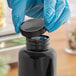 A hand in a blue glove opening a black plastic bottle with a black ribbed cap.