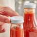 A white metal lid with a plastisol liner being placed on a glass bottle filled with red sauce.