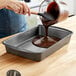 A woman pouring chocolate sauce into a Wilton rectangular cake pan.