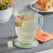 A glass cup of Dona herbal tea with a tea bag in it on a table with a pastry.