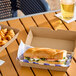 A corrugated clamshell container holding a sandwich, placed on a table next to a box of curly fries and a drink.