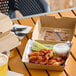 A corrugated clamshell container with a clear window, shown holding chicken wings, celery sticks, and a cup of dipping sauce.