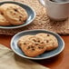 A plate of Otis Spunkmeyer chocolate chip cookies next to a mug of coffee on a table.