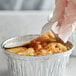 A gloved hand using a spoon to put macaroni in a Baker's Lane foil container.