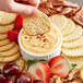 A hand holding a cracker over a bowl of Don's Salads Roasted Garlic and Dijon Cheddar Cheese Spread with crackers and fruit.