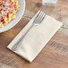 A fork on a Choice Touchstone beige linen feel dinner napkin next to a plate of pasta.