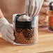 A person in gloves holding a Prepara Evak Fresh Saver clear plastic round food storage container filled with brown balls.