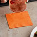 A hand holding a glass with a drink and an orange beverage napkin on a table.