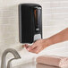 A hand holding a Lavex black manual liquid soap dispenser.