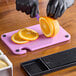 A person using a purple Choice cutting board with black grips to slice an orange.