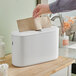 A hand using a Tork white countertop paper towel dispenser to get a brown paper towel.