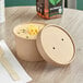 A round Choice kraft paper food container with vented paper lid filled with soup on a table with a wooden spoon.