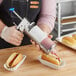 A man using a stainless steel sauce gun to pour ketchup onto a hot dog.