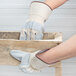 A person wearing Cordova Tuf-Cor white canvas work gloves with heavy side split leather palm coating and rubber cuffs holding a wooden box.