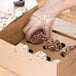 A person wearing a plastic glove placing a chocolate frosted cupcake with sprinkles in a Baker's Mark cupcake box.