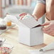 A person's hand opening a Baker's Mark white cupcake box to reveal a cupcake inside.