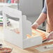 A person wearing gloves cuts a white and purple cake in a Baker's Mark white window cake box.