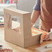 A person in a brown apron and gloves opening a Baker's Mark cake box with a window.