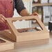 A person holding a Baker's Mark cardboard bakery box with a clear window and donuts inside.