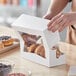 A person's hand opening a white Baker's Mark bakery box to reveal donuts.