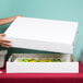 A person holding a white Baker's Mark bakery box with a cake inside.