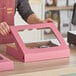 A woman holding a pink Baker's Mark bakery box with a clear window.