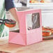 A person holding a chocolate doughnut with sprinkles in a pink Baker's Mark bakery box with a window.