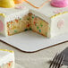 A white frosted cake with pink frosting and colorful sprinkles on a Baker's Mark full sheet cake pad.