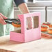 A hand using tongs to pick up a donut with pink sprinkles from a Baker's Mark bakery box with a window.