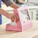 A person opening a Baker's Mark pink bakery box with a clear window on a counter.