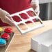 A person holding a white Baker's Mark cupcake box with holes for 6 cupcakes.