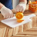 A person using a Choice white polyethylene paddle cutting board with a handle to cut an orange.