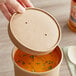 A hand holding a Choice 32 oz. Kraft Paper Soup Container with soup and a vented lid.