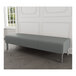 A Lesro Luxe Lounge Series grey fabric bench with steel legs in a room with a white wall.