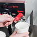 A person using a Grindmaster automatic coffee brewer to fill a white cup with hot water.