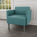 A Lesro Luxe Lounge Patriot Plus chair in teal blue vinyl with steel legs in a lounge area.