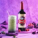 A close-up of a Torani Puremade Galaxy flavoring syrup bottle next to a glass of liquid.