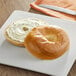 A Just Bagels plain bagel with cream cheese on a plate with a knife.