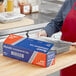 A person in a kitchen wearing a red apron and gloves cuts open a blue and white box of Reynolds Foodservice Heavy-Duty Aluminum Foil.