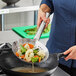 A woman uses an Emperor's Select stainless steel wok skimmer to pour vegetables into a wok.