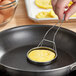 A hand holding an American Metalcraft egg ring with a coil handle over a pan with an egg cooking in it.