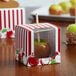A red and white striped Baker's Lane candy apple box with a window and a green candy apple inside.