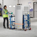 A woman wearing a yellow safety vest and gloves uses a Lavex hydraulic steel equipment mover to push a large grey box.