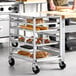 A metal undercounter bun and sheet pan rack with a stainless steel top and side channel, holding trays of baked goods.