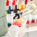A bottle of clear syrup being poured onto a cup of shaved ice, with other colorful snow cones in the background.