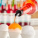 A bottle of orange snow cone syrup being poured over a cup of shaved ice.