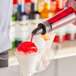 A bottle of red syrup being poured onto a snow cone in a paper cup.