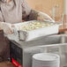 A woman in a chef's uniform using a Vollrath stainless steel steam table pan to prepare food.