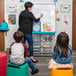 A woman teaching children in a classroom using a Luxor white steel mobile modular teacher dry erase easel.