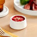 A white porcelain sauce cup filled with red sauce on a wooden table.